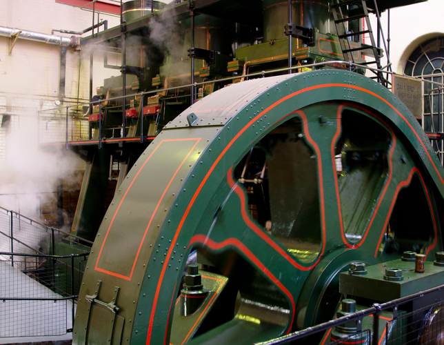 A photograph of a large engine inside a room. There is a sloped viewing platform in front of the engine. The engine is surrounded by a black barrier. Steam can be seen coming out of the engine.  
