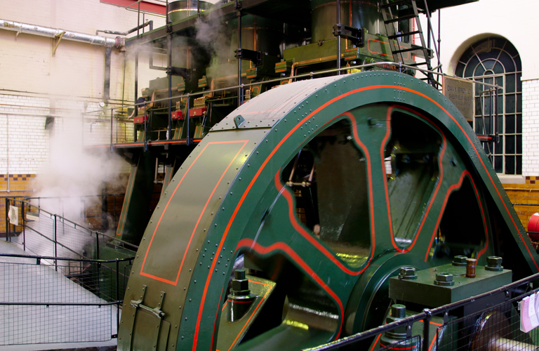 A photograph of a large engine inside a room. There is a sloped viewing platform in front of the engine. The engine is surrounded by a black barrier. Steam can be seen coming out of the engine.  