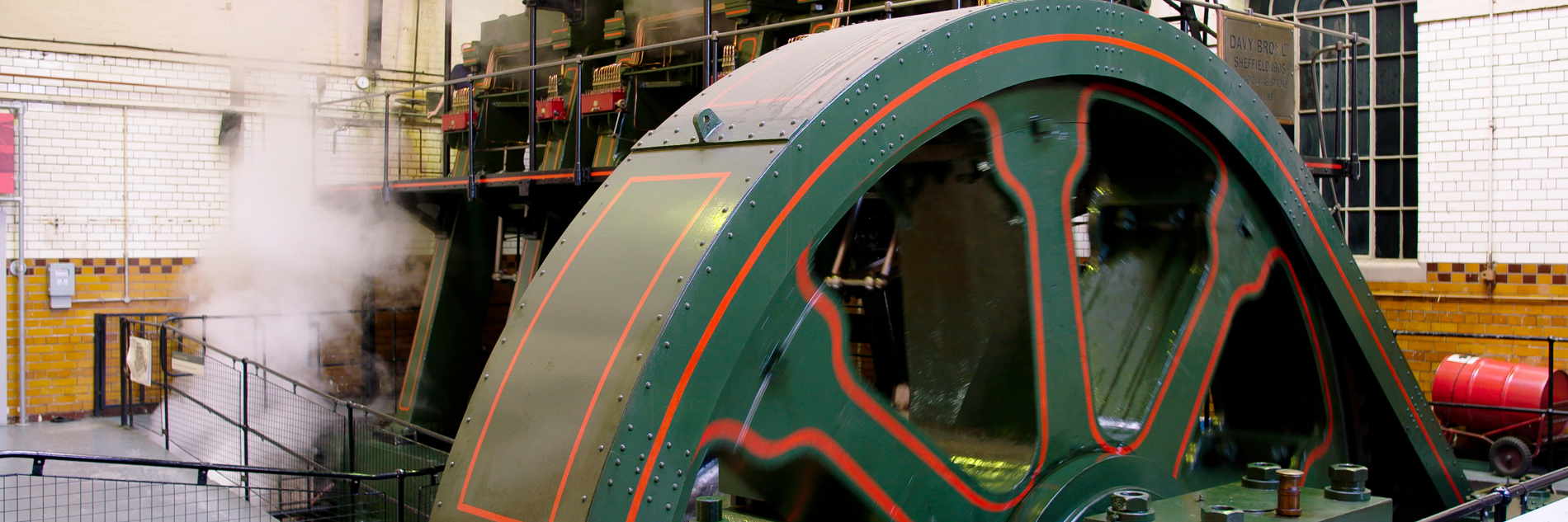 A photograph of a large engine inside a room. There is a sloped viewing platform in front of the engine. The engine is surrounded by a black barrier. Steam can be seen coming out of the engine.  