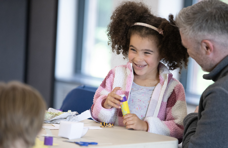 A photograph of a young girl smiling while holding a glue stick. An adult man sits to her right and the table in front of them is full of crafting materials