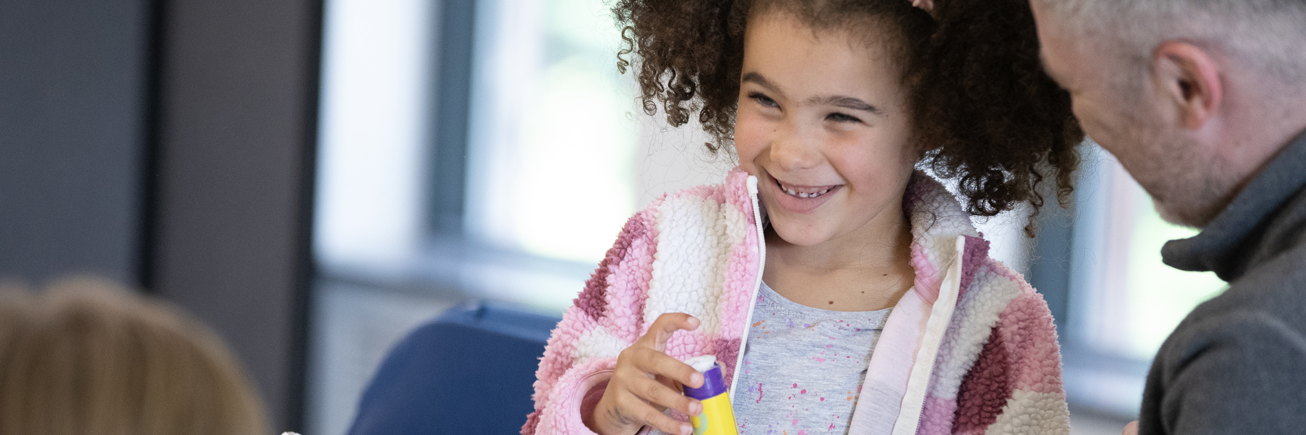 A photograph of a young girl smiling while holding a glue stick. An adult man sits to her right and the table in front of them is full of crafting materials