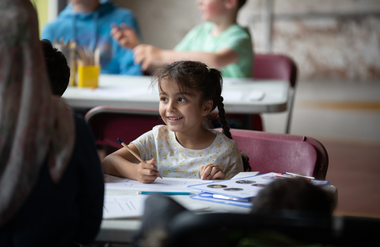 A smiling girl with two long plaits is sat at a table with a pencil in her hand. There is a clipboard with printed materials on the table next to her.