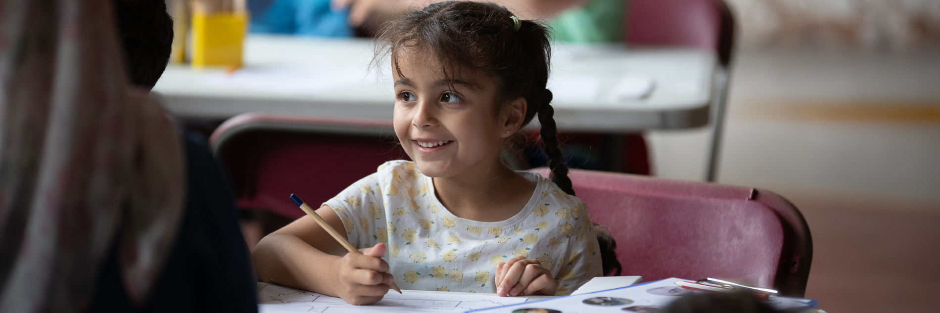A smiling girl with two long plaits is sat at a table with a pencil in her hand. There is a clipboard with printed materials on the table next to her.
