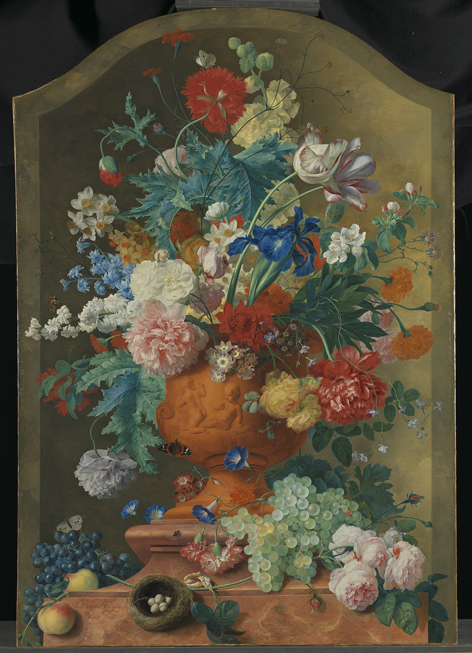 An arrangement of blue, pink, white and red flowers in an orange vase. The vase is surrounded by fruit including black and green grapes and a bird's nest with eggs in it.