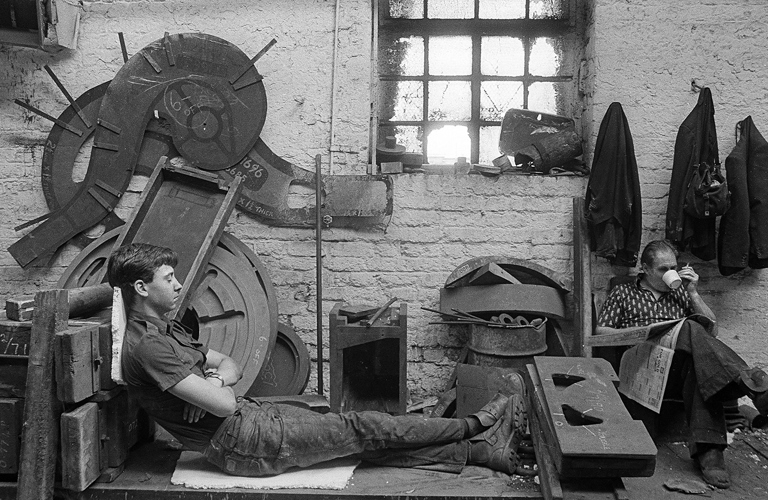 A black and white photograph of two adults in a workshop.  One adult is leaning against equipment with their eyes closed and the other adult is sitting in a chair, reading a newspaper and drinking out of a cup whilst holding a cigarette. 