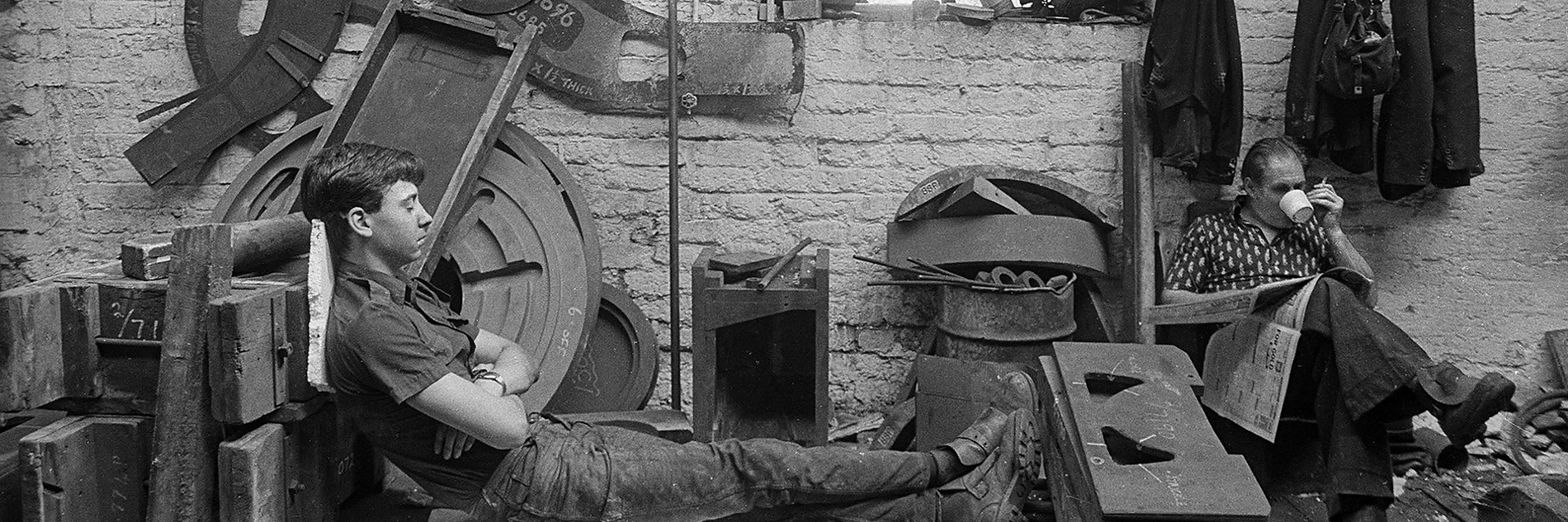 A black and white photograph of two adults in a workshop.  One adult is leaning against equipment with their eyes closed and the other adult is sitting in a chair, reading a newspaper and drinking out of a cup whilst holding a cigarette. 