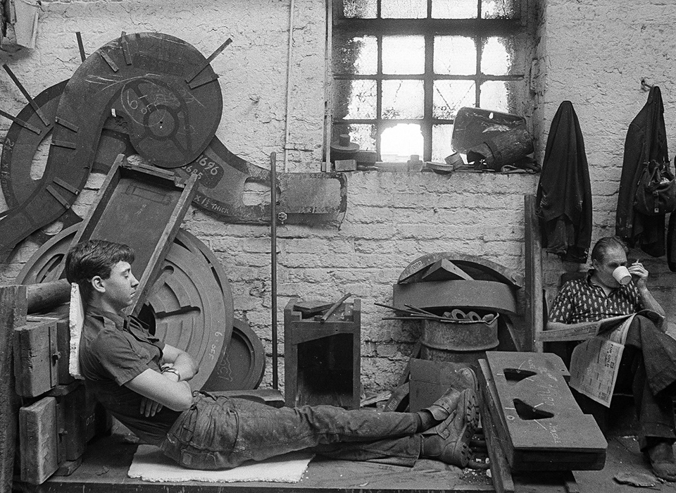 A black and white photograph of two adults in a workshop.  One adult is leaning against equipment with their eyes closed and the other adult is sitting in a chair, reading a newspaper and drinking out of a cup whilst holding a cigarette. 