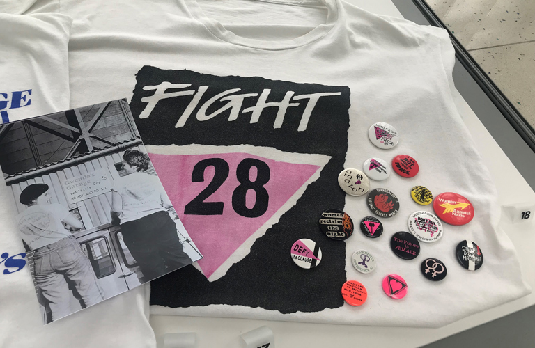 Part of a display in a museum case showing a white t-shirt with a black and pink graphic and the words 'Fight 28'. To the left is a black and white photograph of two female mechanics wearing t-shirts with the Gwenda's Garage logo. To the right is a selection of colourful protest badges.