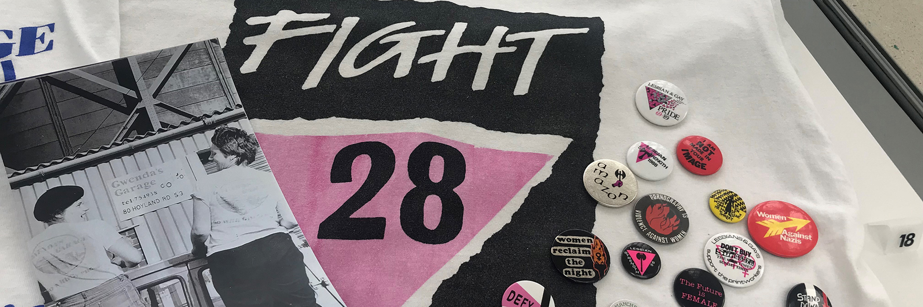 Part of a display in a museum case showing a white t-shirt with a black and pink graphic and the words 'Fight 28'. To the left is a black and white photograph of two female mechanics wearing t-shirts with the Gwenda's Garage logo. To the right is a selection of colourful protest badges.