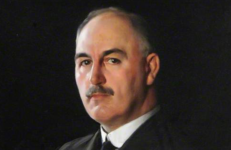 A portrait painting of Dr J.G Graves at around 77 years old. He looks out from the canvas directly with a serious expression and has short, grey hair, a neat moustache and he's wearing a dark suit againist a plain, black background. 