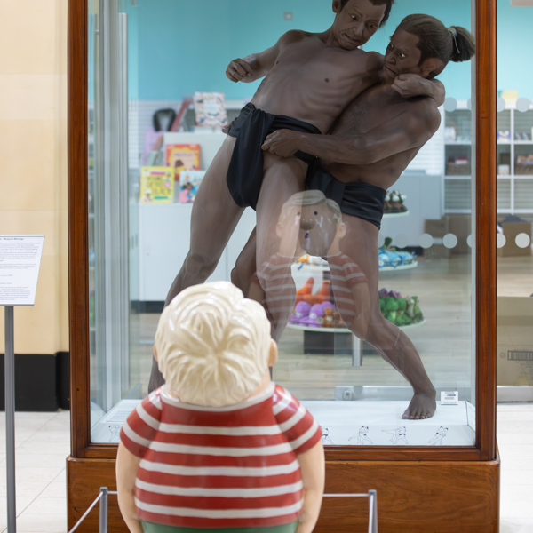 A cartoon sculpture of a child looks up at a lifelike historic sculpture of Japanese wrestlers in a glass and wood case