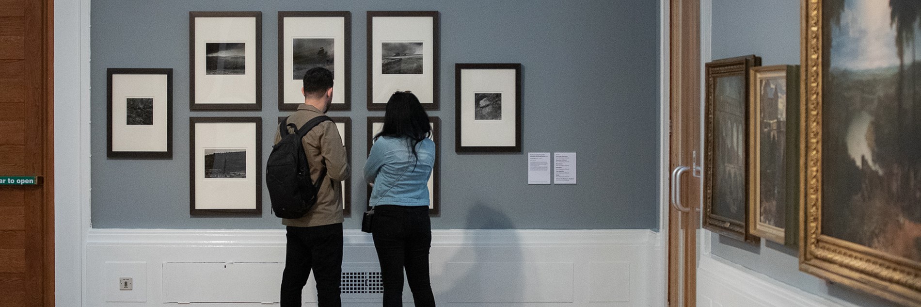 A photograph of two gallery visitors looking at a display of seven black and white framed photographs displayed on a grey wall.