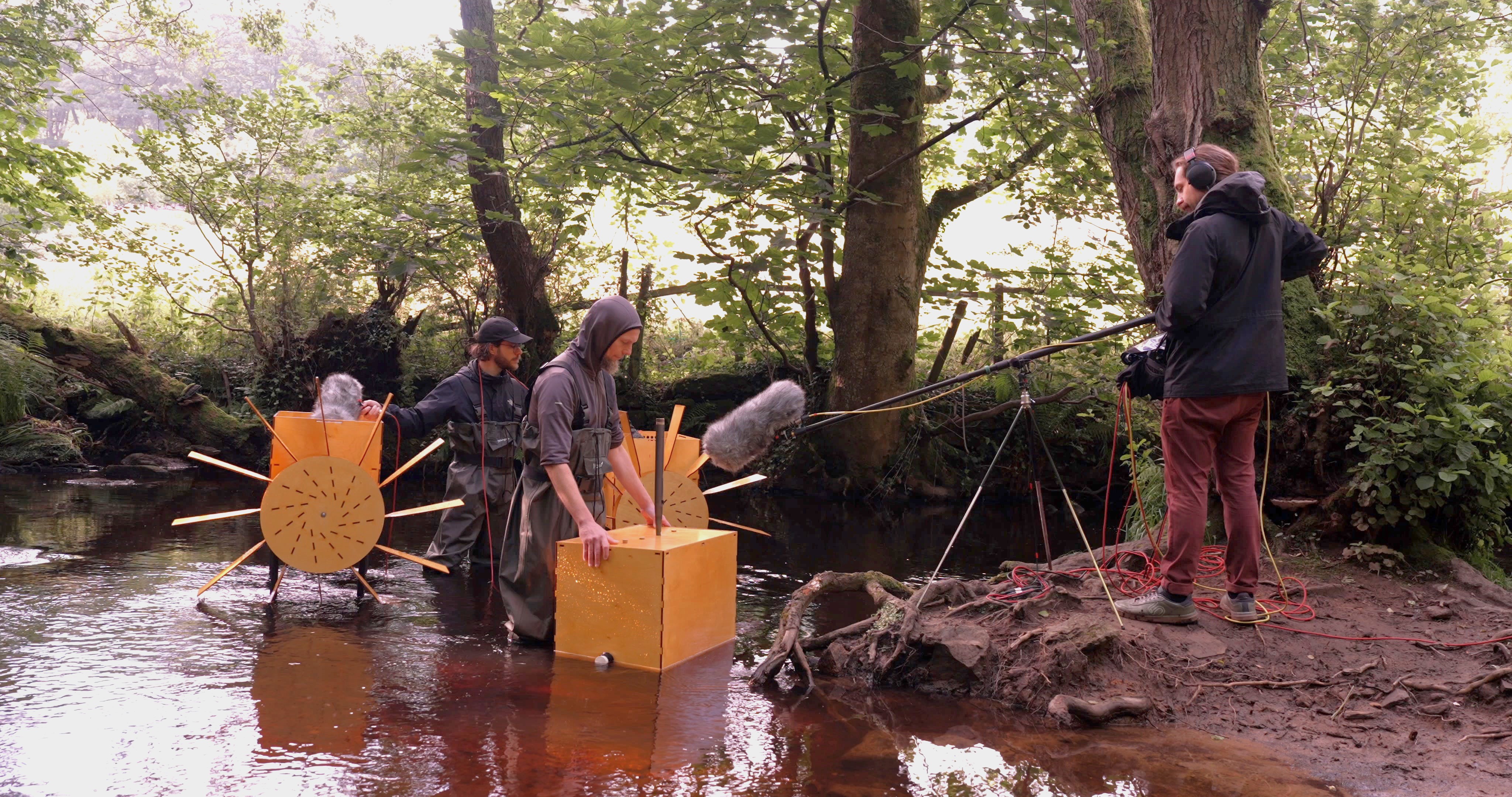 Two musicians playing water-powered mechanical musical instruments on the banks of the River Rivelin. They're wearing waders while someone on the banks of the river records the music using a long-arm microphone over the water.