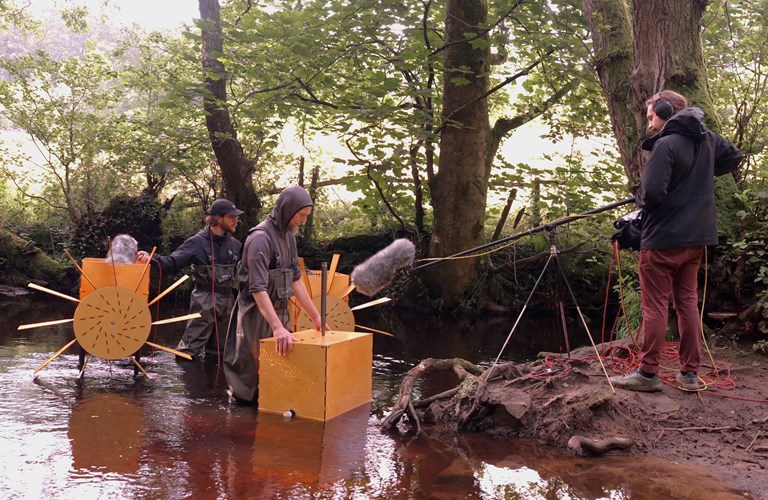 Two musicians playing water-powered mechanical musical instruments on the banks of the River Rivelin. They're wearing waders while someone on the banks of the river records the music using a long-arm microphone over the water.