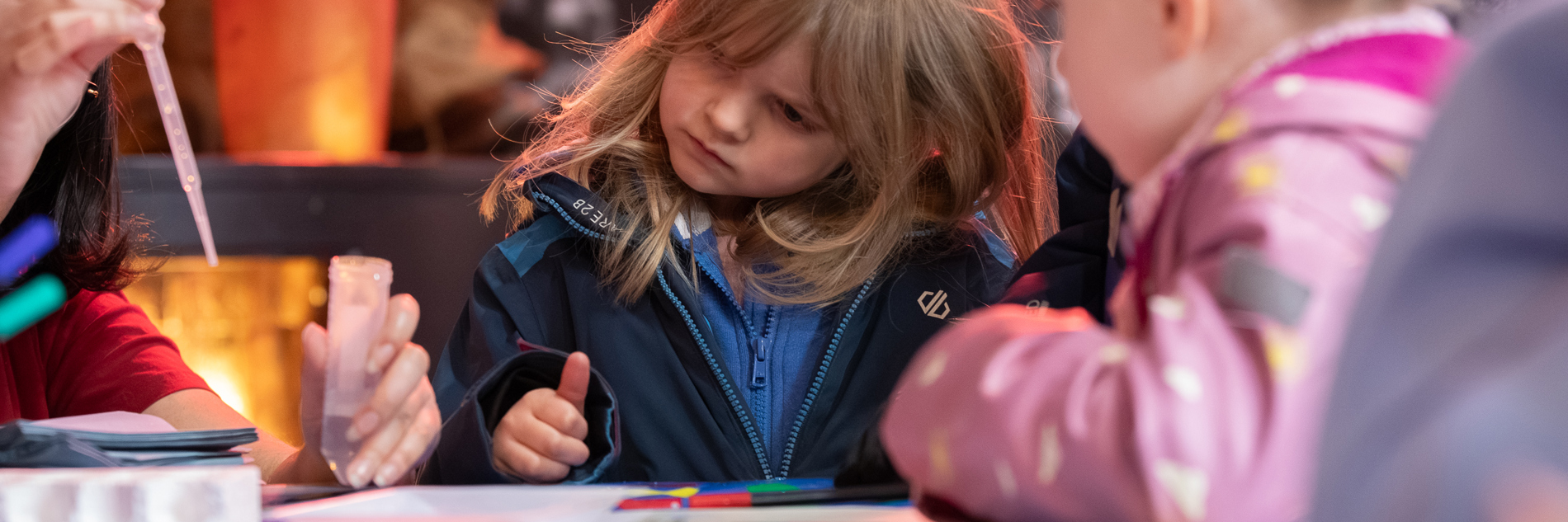 A photograph of two girls watching an adult do an experiment using a test tube and a pipette.