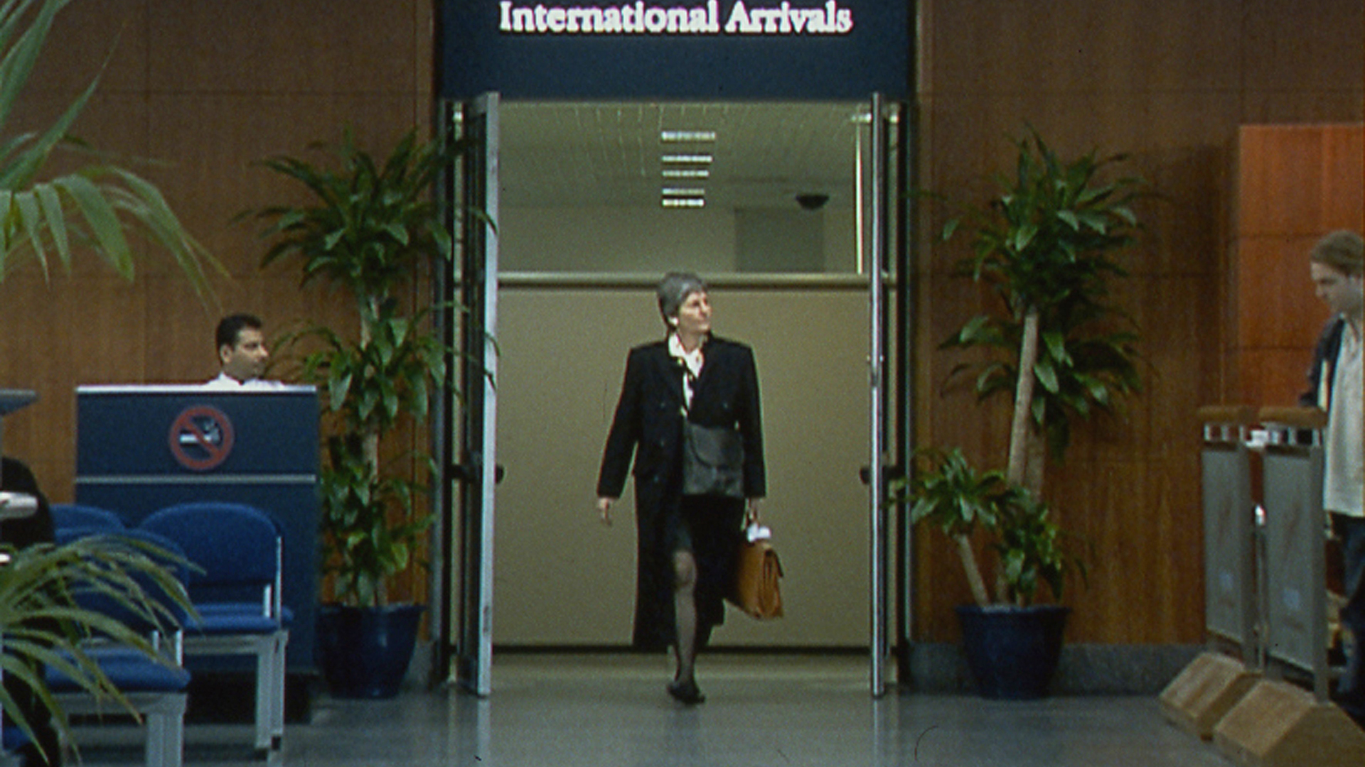 A video still of a smartly dressed person walking through a doorway marked "International Arrivals" at an airport terminal. To the left of the doorway there is a a person is sitting at a desk with a large "No Smoking Sign" on the front of it and chairs in front and to the right of the doorway a person is waiting behind a barrier.  