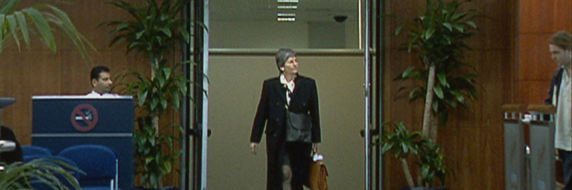 A video still of a smartly dressed person walking through a doorway marked "International Arrivals" at an airport terminal. To the left of the doorway there is a a person is sitting at a desk with a large "No Smoking Sign" on the front of it and chairs in front and to the right of the doorway a person is waiting behind a barrier.  