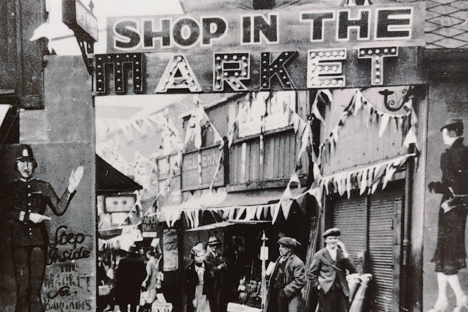 A black and white photograph from the 1930s of a Rag N Tag Market. The photograph is taken at the entrance, with the words 'Shop in the market' shown in large letters at the top of a rectangular entryway. Through the entryway you can see an alley with lots of bunting on the shop fronts and people walking around underneath. A drawing of a policeman is shown on the left side of the entryway, encouraging people to step inside.