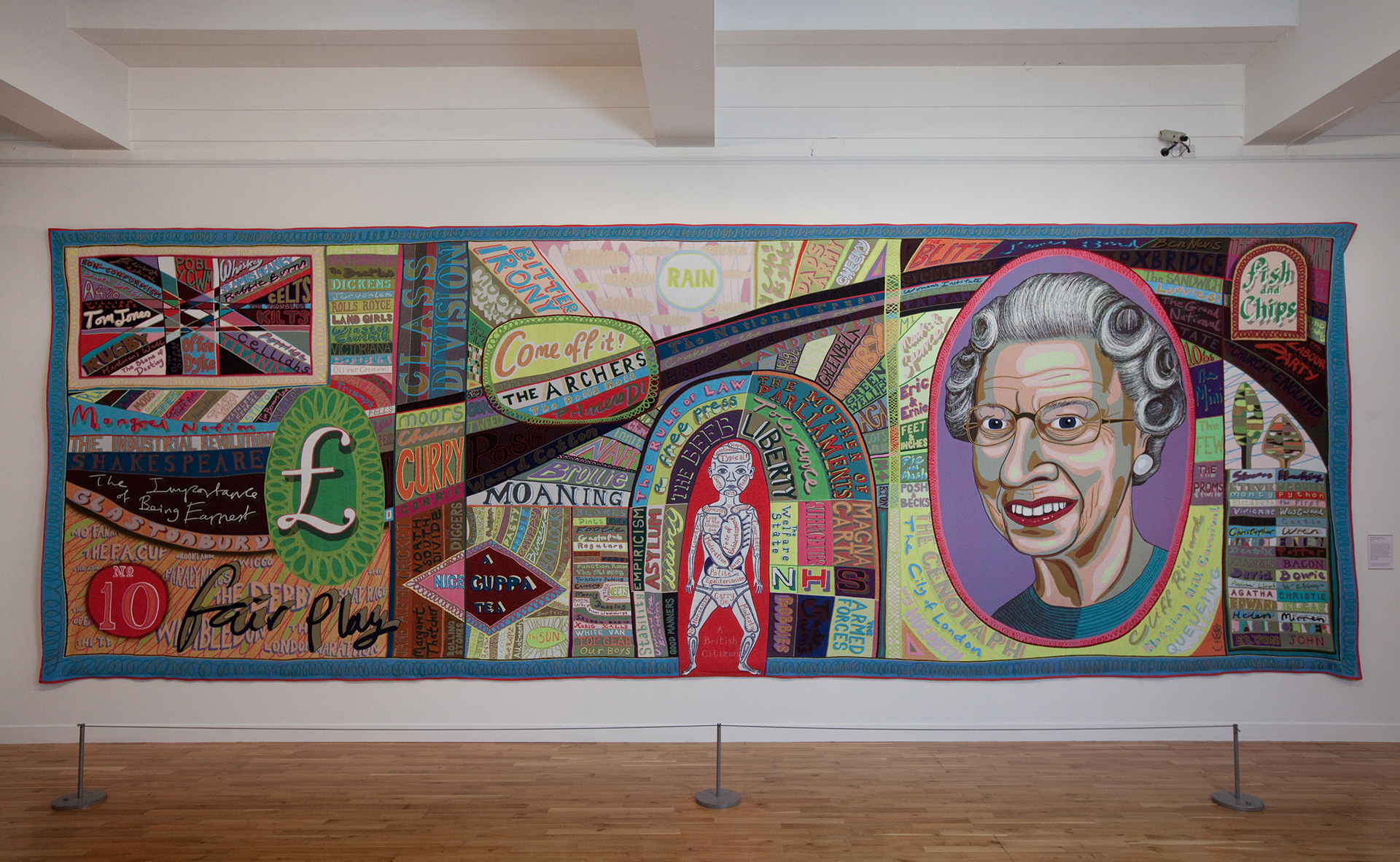 A multi-coloured tapestry in a long landscape format incorporating a huge variety of text and images on the theme of ‘Britishness’. An image of Queen Elizabeth II and the pound symbol feature prominently.			
