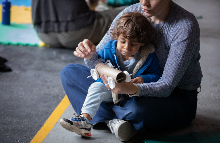 A photograph of a young boy sat on an adults lap. The adult is holding a car made out of a cardboard tube, sticks and bottle tops. They are both sat on the floor