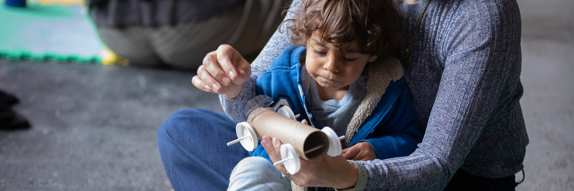 A photograph of a young boy sat on an adults lap. The adult is holding a car made out of a cardboard tube, sticks and bottle tops. They are both sat on the floor