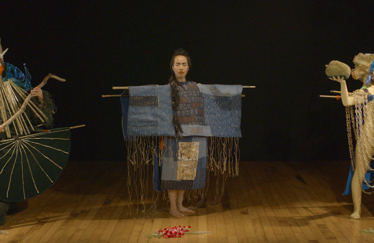 Three performers stand on a wooden floor, each wearing differently constructed costumes. Flowers are laid on the floor in front of them. 