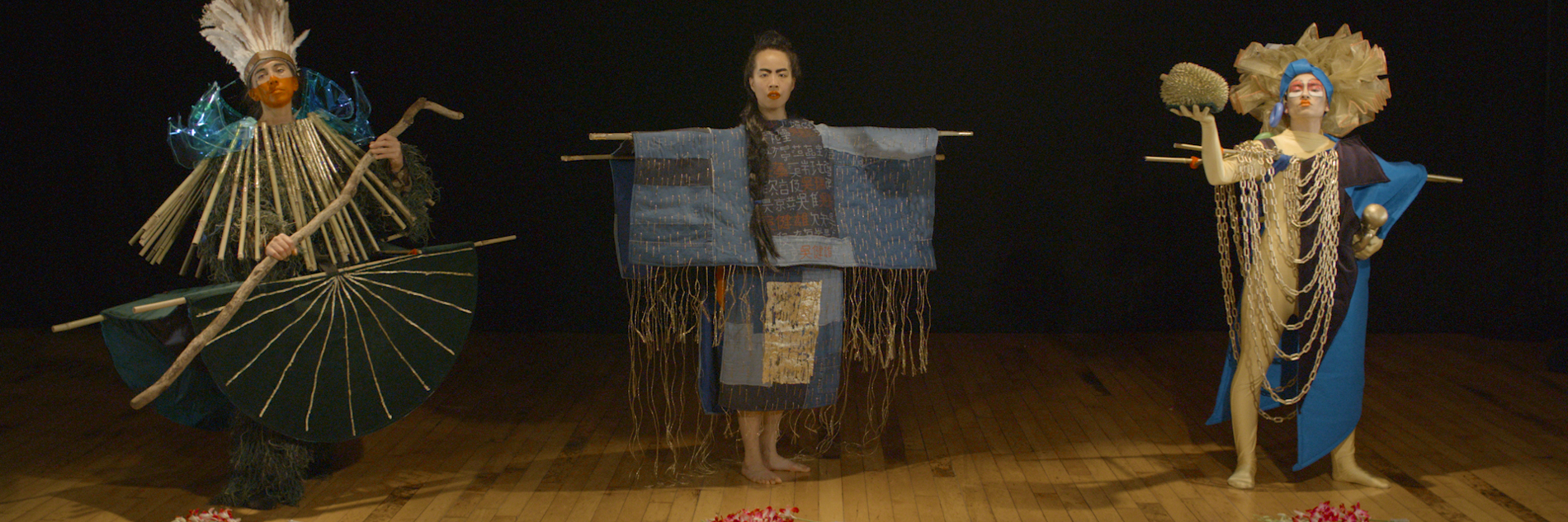Three performers stand on a wooden floor, each wearing differently constructed costumes. Flowers are laid on the floor in front of them. 