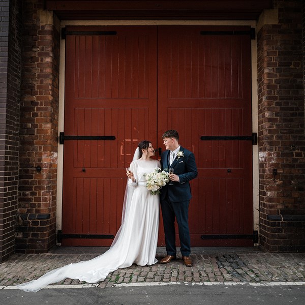 A bride in a wedding dress and a groom in a suit pose in front of a large red doors of an industrial building. 