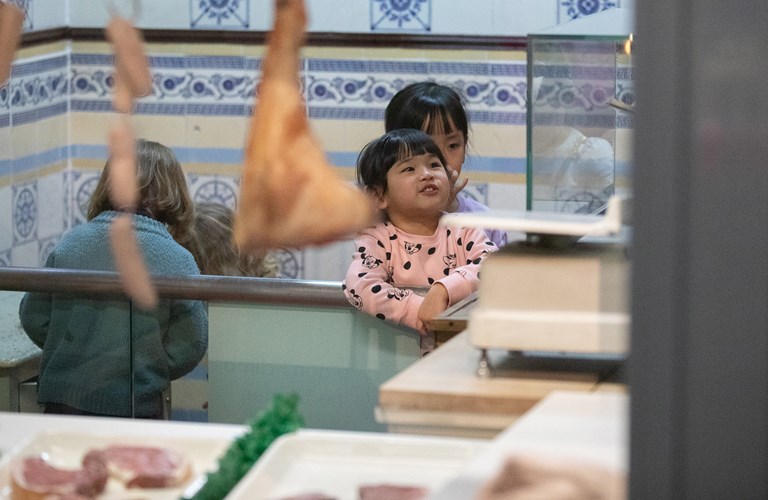 Four young children in the museum's shop recreation, one smiling and leaning against a glass counter. There are plastic meat products in trays in the foreground with sausages and legs of meat hanging down with a background of  blue and white patterned tiles.