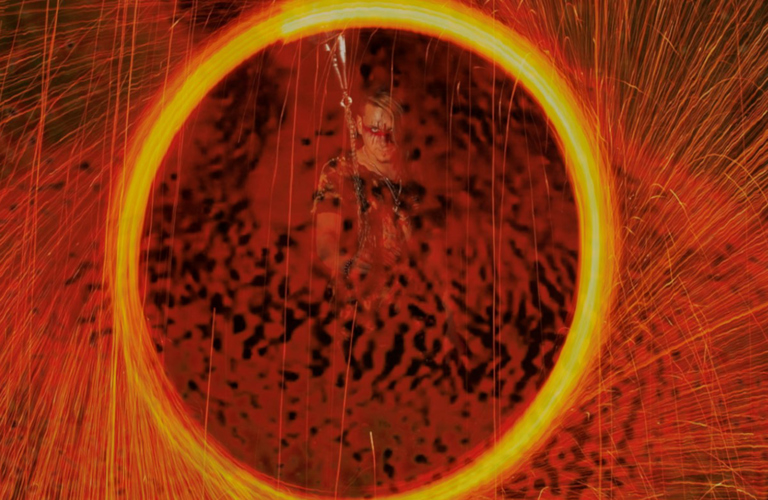 A photograph of a large yellow circle made up of yellow sparks with sparks surrounding the circle. Inside the circle an adult wearing red and black face paint looks directly at the camera.  