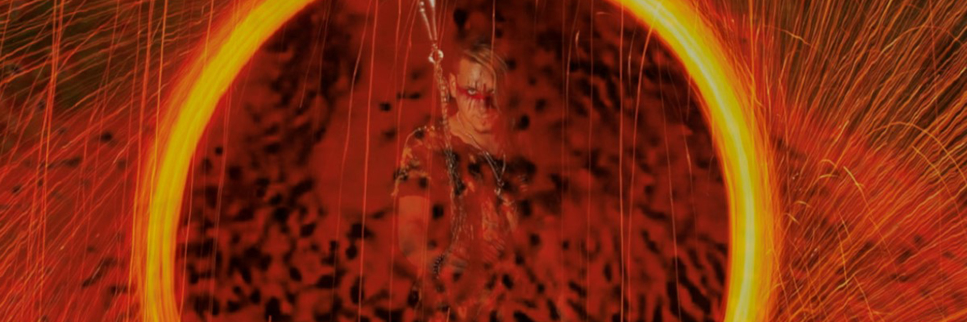 A photograph of a large yellow circle made up of yellow sparks with sparks surrounding the circle. Inside the circle an adult wearing red and black face paint looks directly at the camera.  