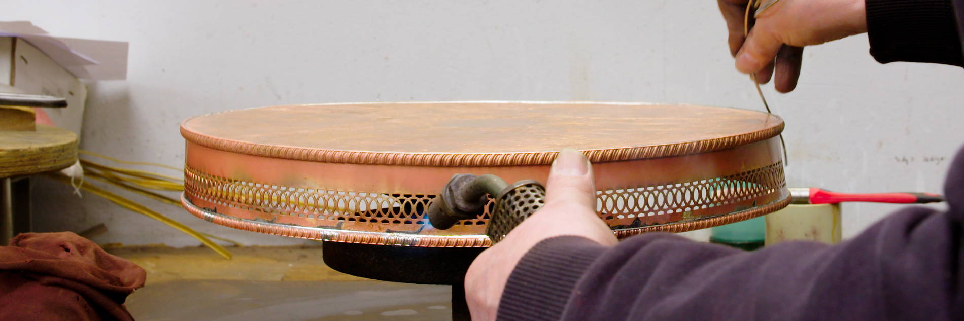 A photograph of arms and hands of a person holding a blow torch towards a circular copper object on a desk. Other objects are also on the desk including a cloth, pot and paintbrush.