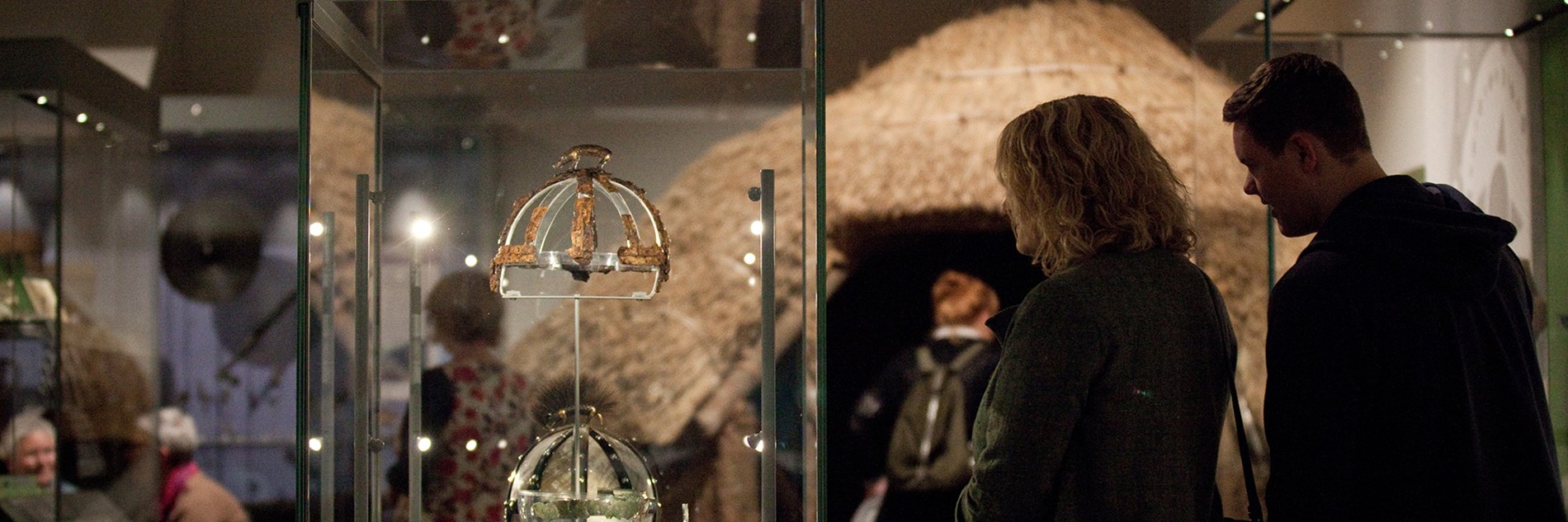 The Beneath Your Feet gallery, with two people looking at the remains of an Anglo-Saxon boar-crested helmet and a roundhouse in the background.