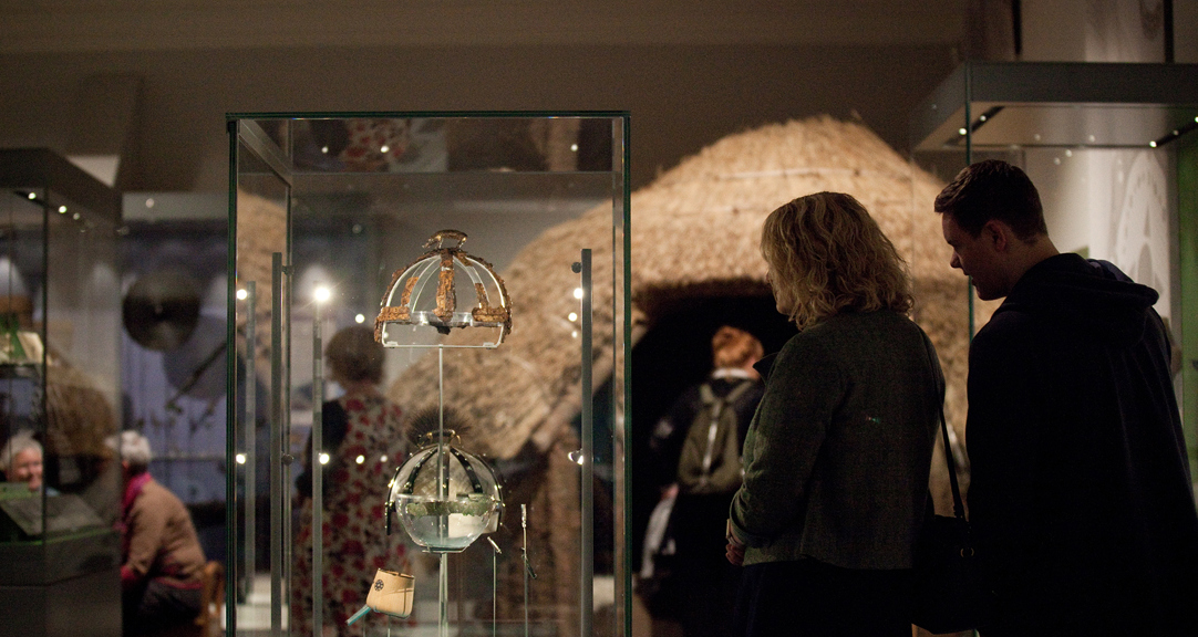 The Beneath Your Feet gallery, with two people looking at the remains of an Anglo-Saxon boar-crested helmet and a roundhouse in the background.