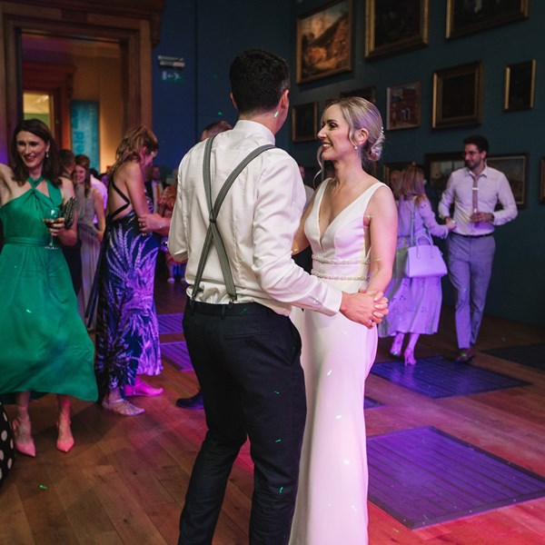 A bride, groom and wedding guests dance in a gallery. the room is lit by coloured lighting and pictures in frames cover the walls.
