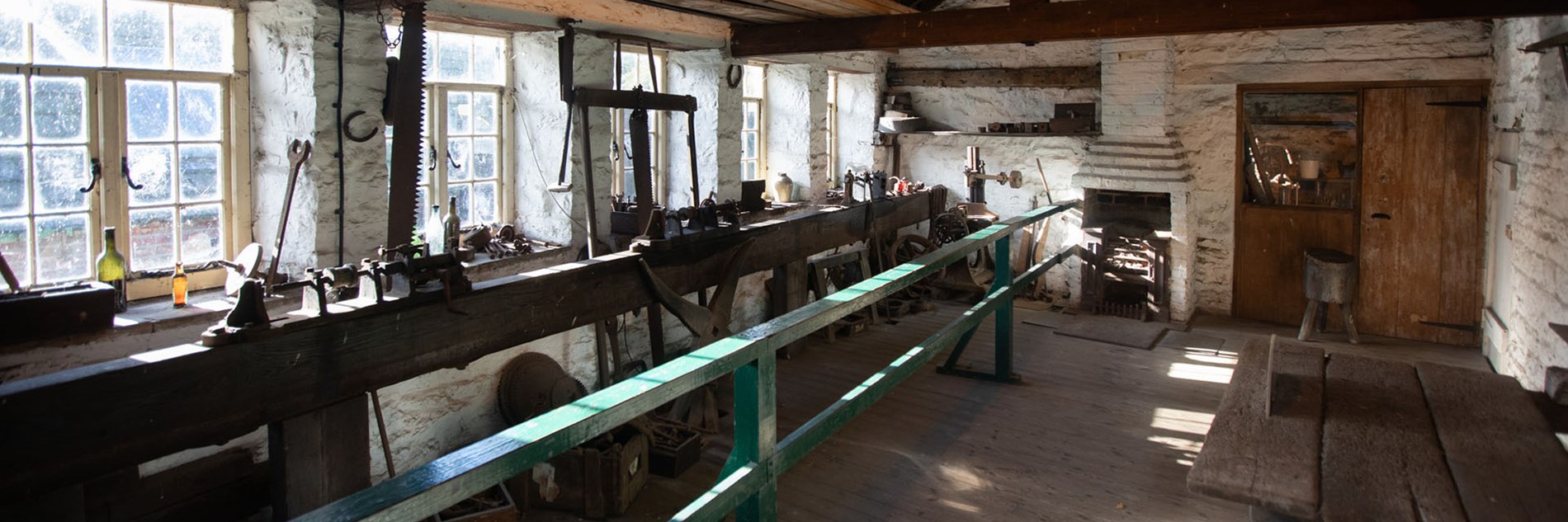 Looking along an old whitewashed workshop with three boring machines mounted on benching backlit by windows. An open fireplace and chimney breast are at the end of the room.