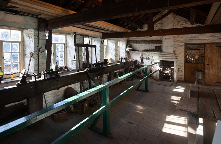 Looking along an old whitewashed workshop with three boring machines mounted on benching backlit by windows. An open fireplace and chimney breast are at the end of the room.