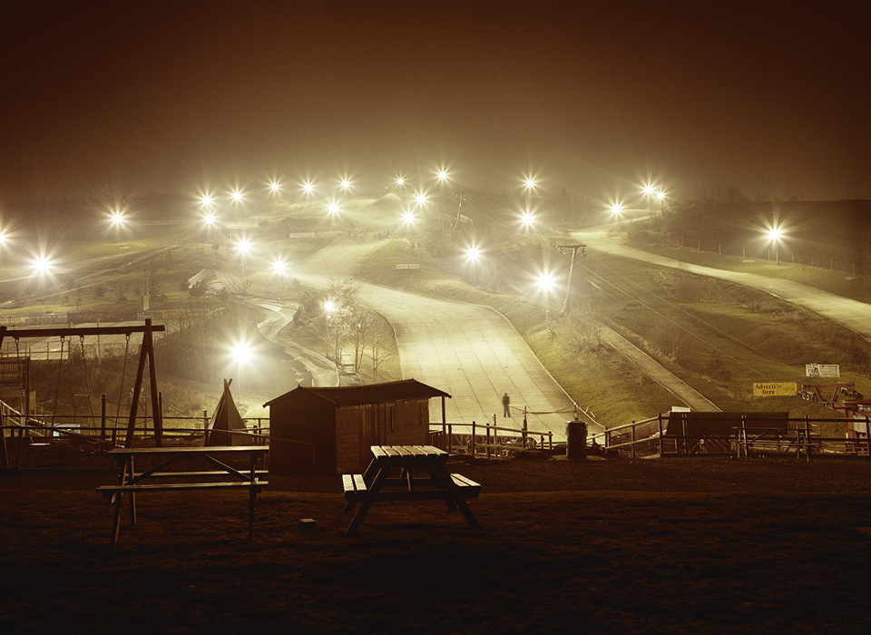 A photograph of a ski slope in Sheffield. There are lots of lightpoles, all with their lights shining brightly, creating a starburst effect in the cameras lens. A few different ski slopes are visible between the lights. A playground and picnic benches are visible in the foreground.