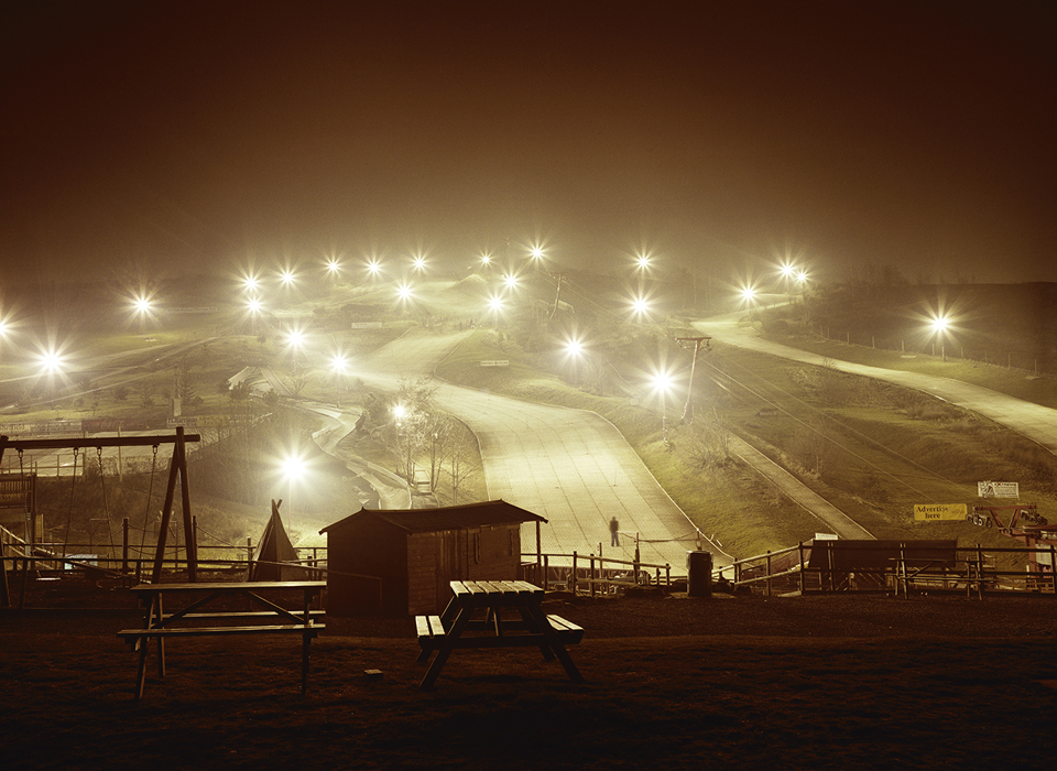 A photograph of a ski slope in Sheffield. There are lots of lightpoles, all with their lights shining brightly, creating a starburst effect in the cameras lens. A few different ski slopes are visible between the lights. A playground and picnic benches are visible in the foreground.