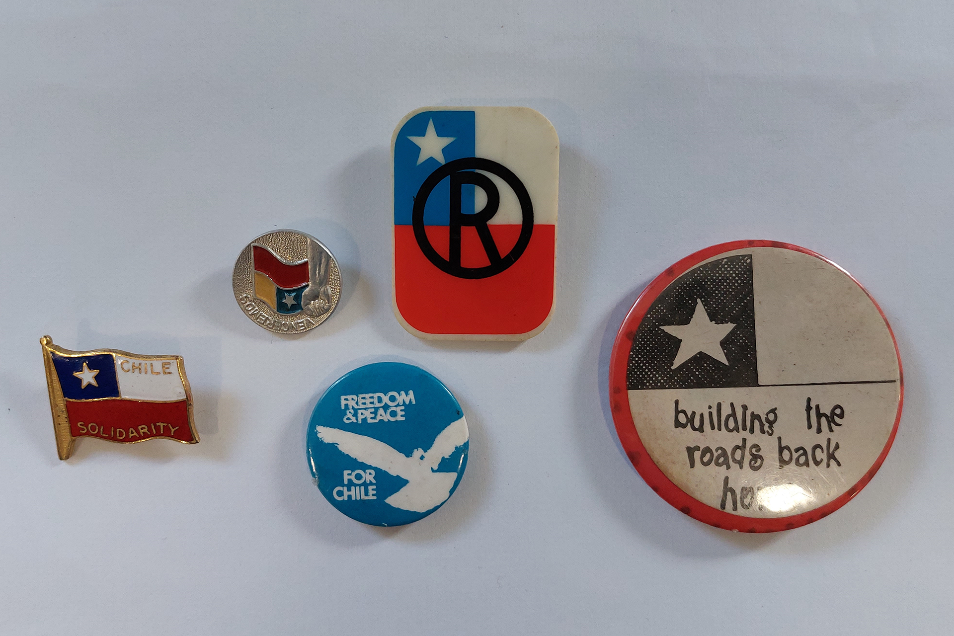 A photograph showing five Chilean badges photographed against a white background. Four feature the flag of Chile, the fifth shows a white bird against a blue background with the words 'Freedom & Peace for Chile'.