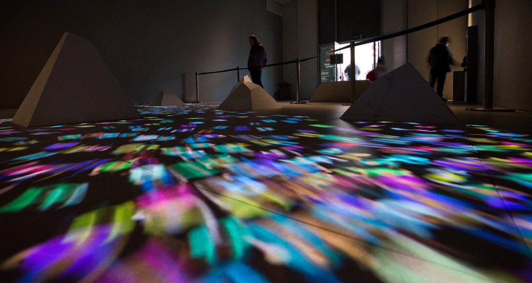 Gallery floor with truncated pyramidal shapes and a stream of multi-coloured lights. In the background there is a barrier, several adults and the gallery door. 