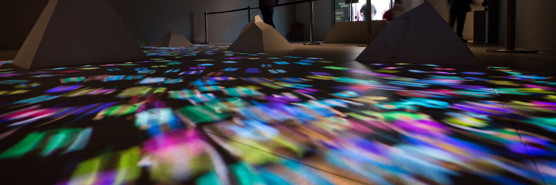 Gallery floor with truncated pyramidal shapes and a stream of multi-coloured lights. In the background there is a barrier, several adults and the gallery door. 