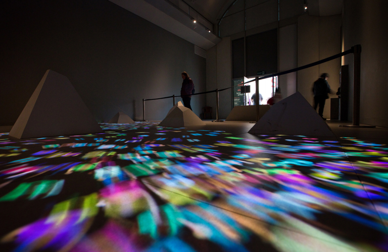 Gallery floor with truncated pyramidal shapes and a stream of multi-coloured lights. In the background there is a barrier, several adults and the gallery door. 