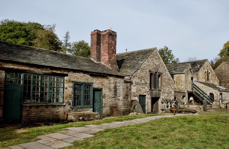 A landscape photograph of one of the old stone cottages at Abbeydale Industrial Hamlet. A chimney stack is in the centre of the image, and grass and a path are shown in front of the cottage.