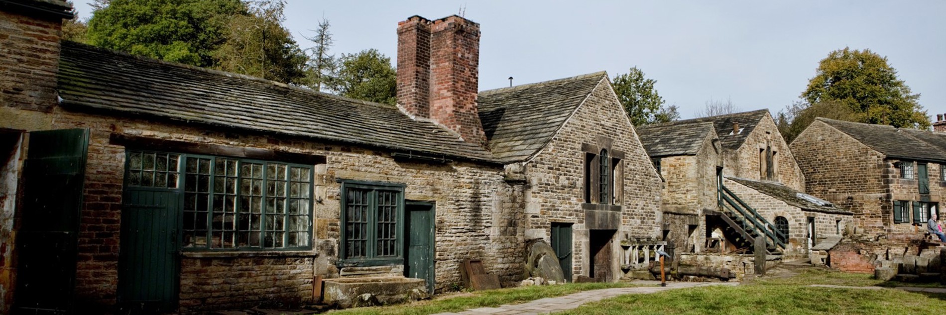 A landscape photograph of one of the old stone cottages at Abbeydale Industrial Hamlet. A chimney stack is in the centre of the image, and grass and a path are shown in front of the cottage.
