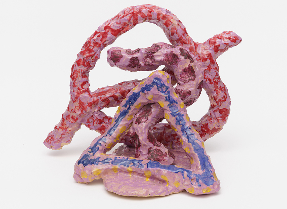  A photograph of Hoyland's work Thupelo Memory, shown against a white background. A pink, yellow and blue painted triangle shape rests a top a pink, oval shaped base. A pink curved shape rests behind it, snaking up to the top of the triangle. Behind that is a large pink circle shape, with a line through it. All are decorated with splodges of red and burgundy paint.