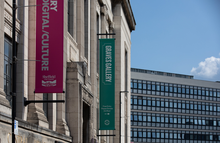 A photograph of the exterior of Graves Gallery on a sunny day, a pink and green banner are running horizontally down the building. A glass walled office building is visible in the background.