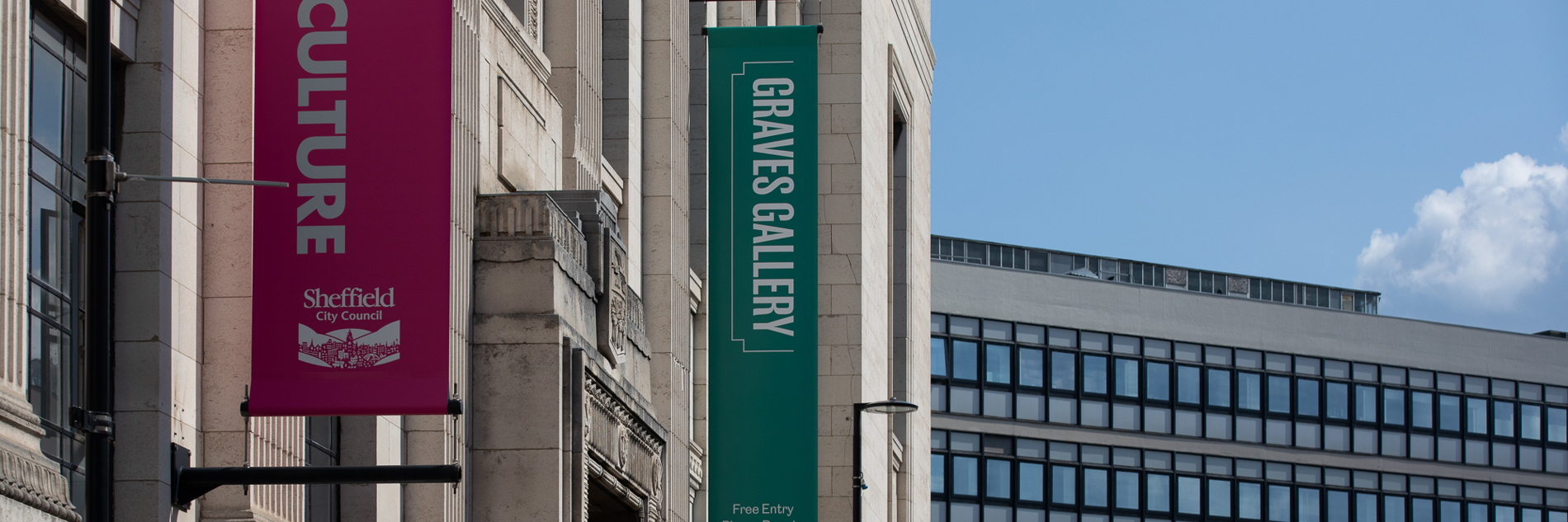 A photograph of the exterior of Graves Gallery on a sunny day, a pink and green banner are running horizontally down the building. A glass walled office building is visible in the background.