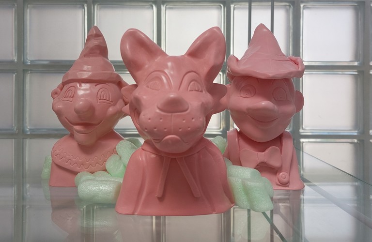 An image of three pink sculptural works presented in a glass display cabinet. On the right there is a clown, middle is an animal wolf and on the left is an interpretation of Pinocchio, all three are busts. There are styrofoam packing peanuts inbetween the pieces.