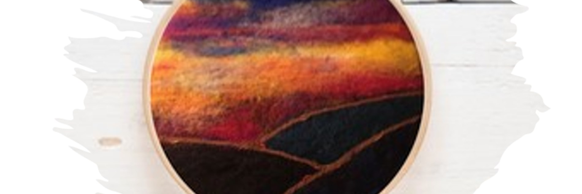Photograph of a needle-felted landscape in an embroidery hoop. The image features dark coloured hills with a red, orange and dark blue sky.